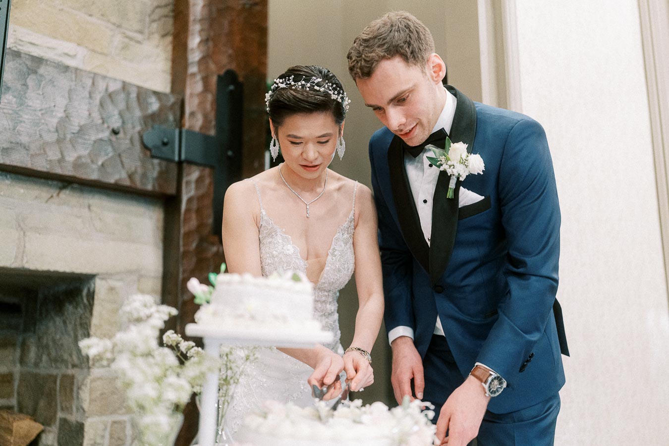 Bride and groom cutting a wedding cake together in a beautifully decorated venue, with the bride in a white gown and the groom in a blue suit.