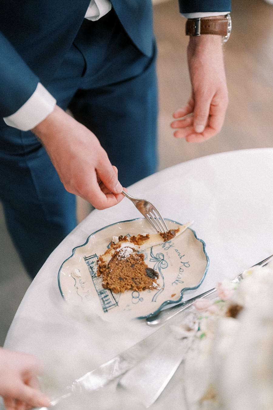 A person in a blue suit cutting a piece of cake on a decorative plate at a wedding reception.