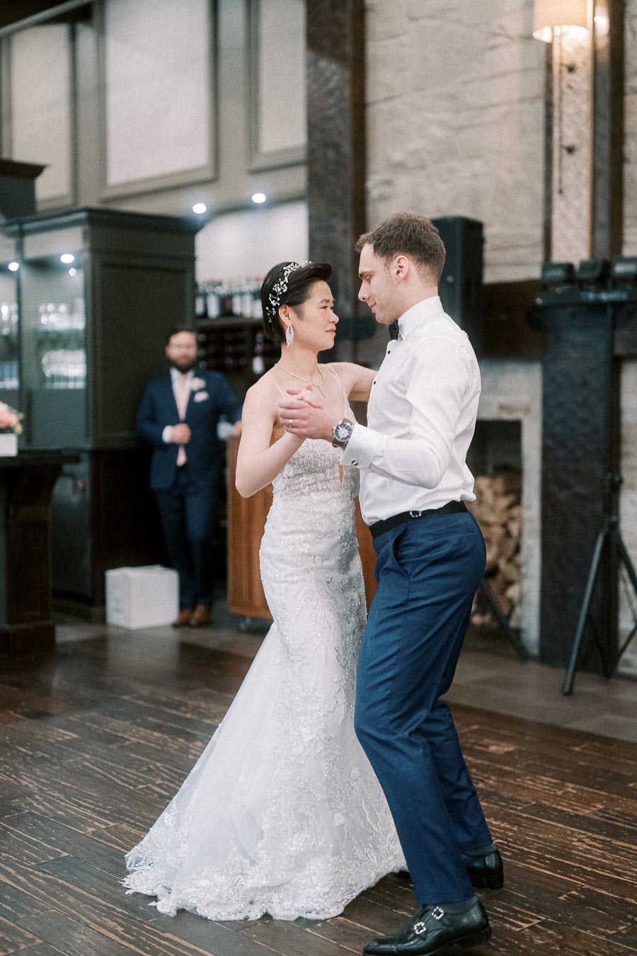 Bride and groom sharing their first dance at a wedding reception, captured in a warmly lit, elegant venue with a stone fireplace as the backdrop.