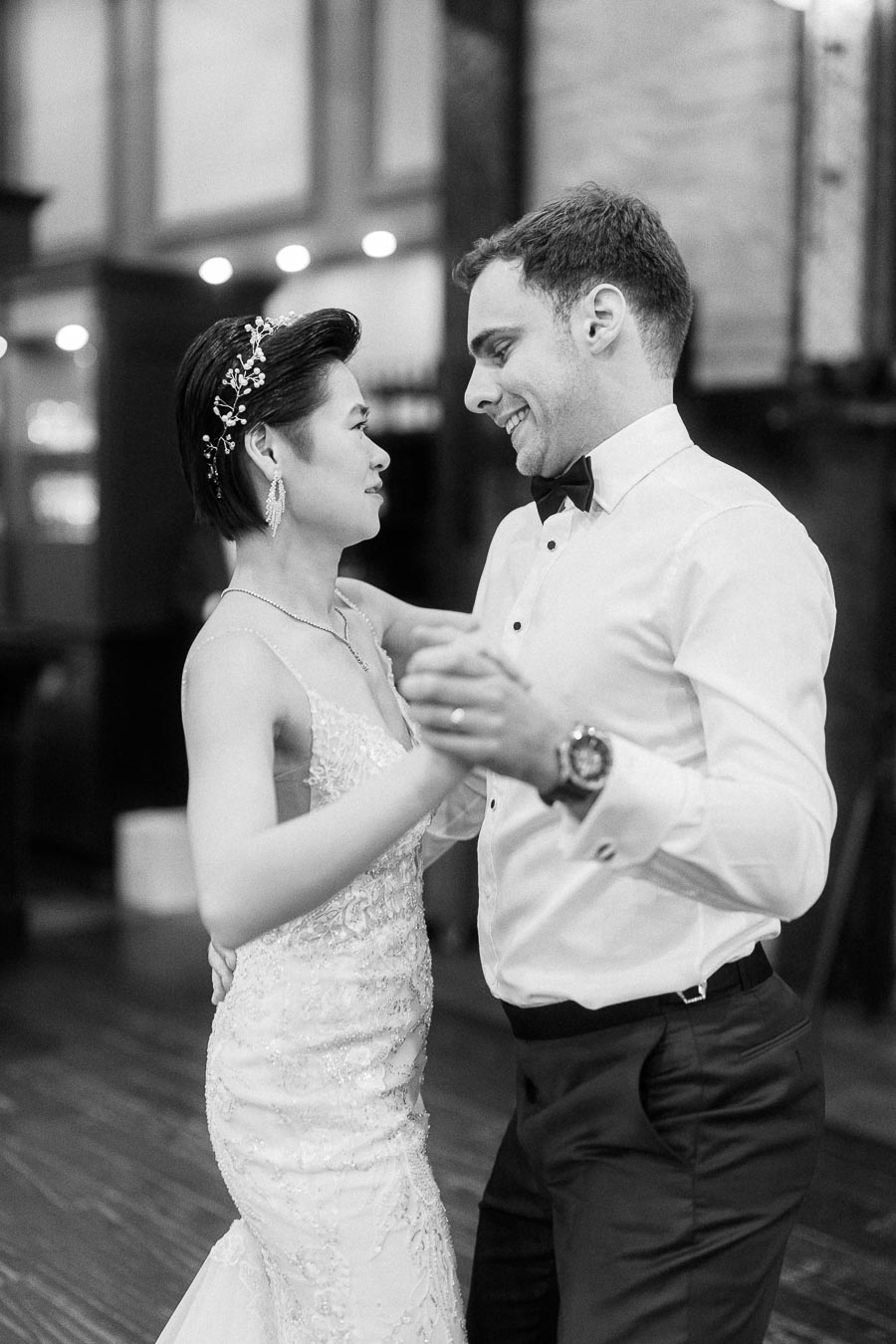 A couple enjoying their first dance on their wedding day, with the bride in an elegant lace gown and the groom in a classic white shirt and black bow tie, captured in a candid, joyful moment.