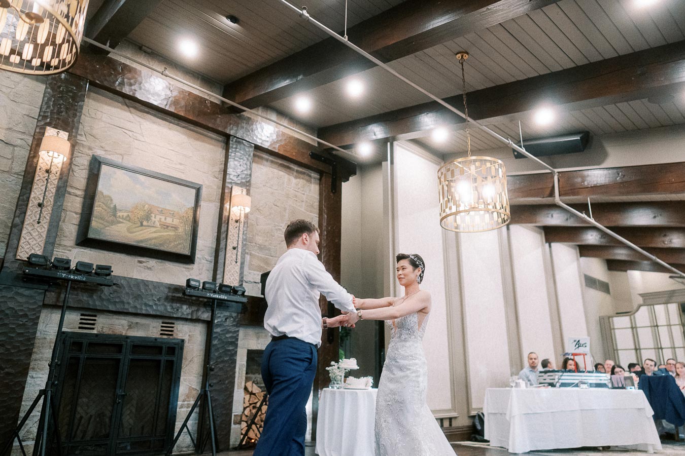 Wedding couple dancing at reception in a beautifully decorated venue with elegant lighting and a stone fireplace backdrop.