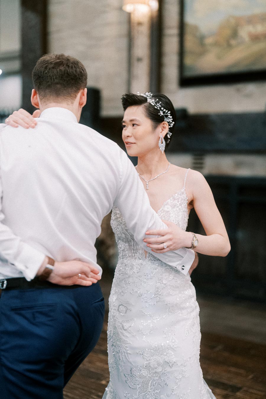 Bride and groom sharing a first dance in an elegant wedding venue, with the bride wearing a lace wedding gown and sparkling accessories, and the groom in a classic formal outfit.