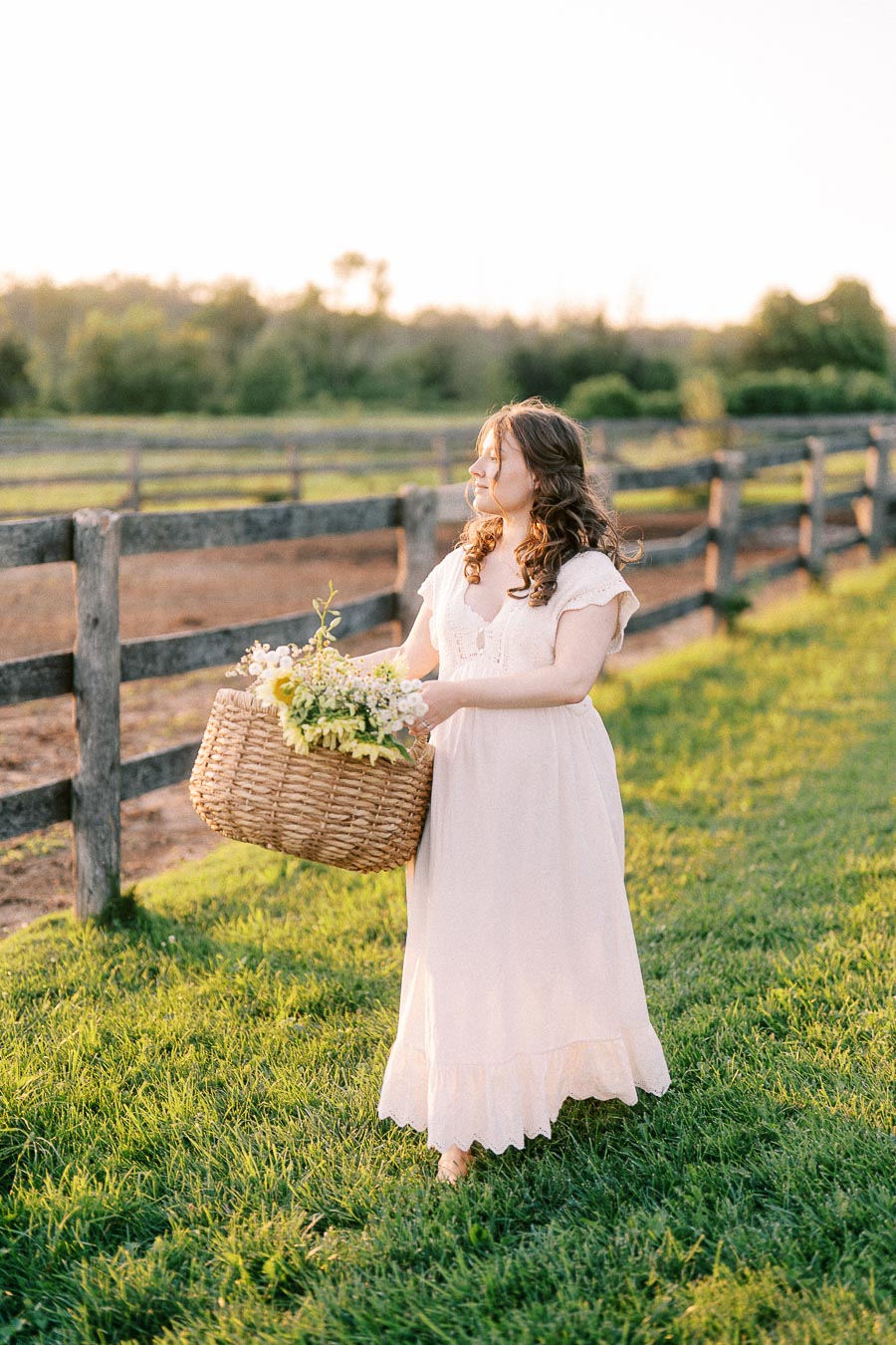 Girl in a white dress holding a flower basket beside a farm fence