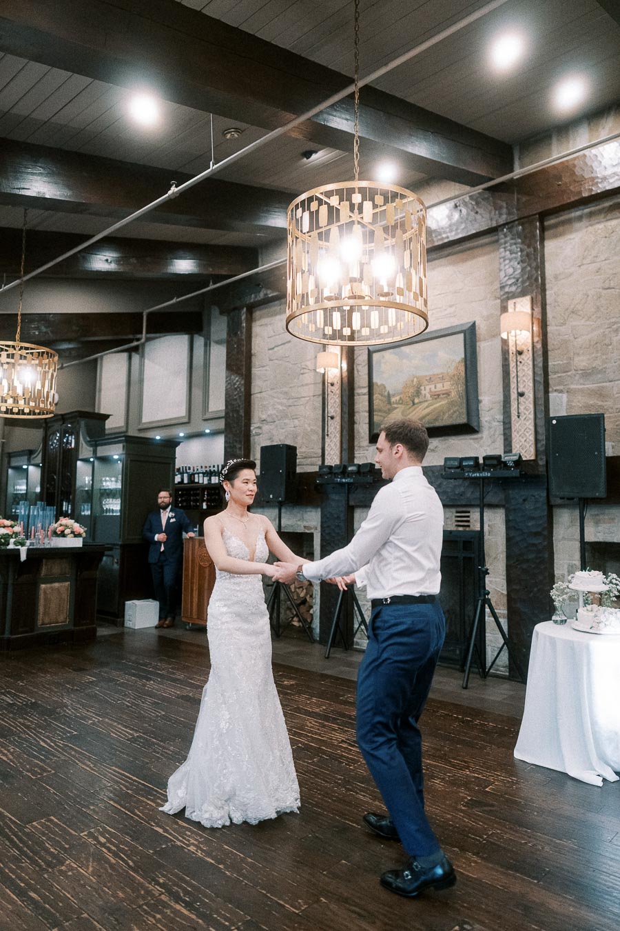 A couple shares a romantic first dance in an elegant wedding venue, featuring rustic wooden beams, warm lighting, and a stone fireplace. The bride wears a stunning lace gown, while the groom is dressed in formal attire.