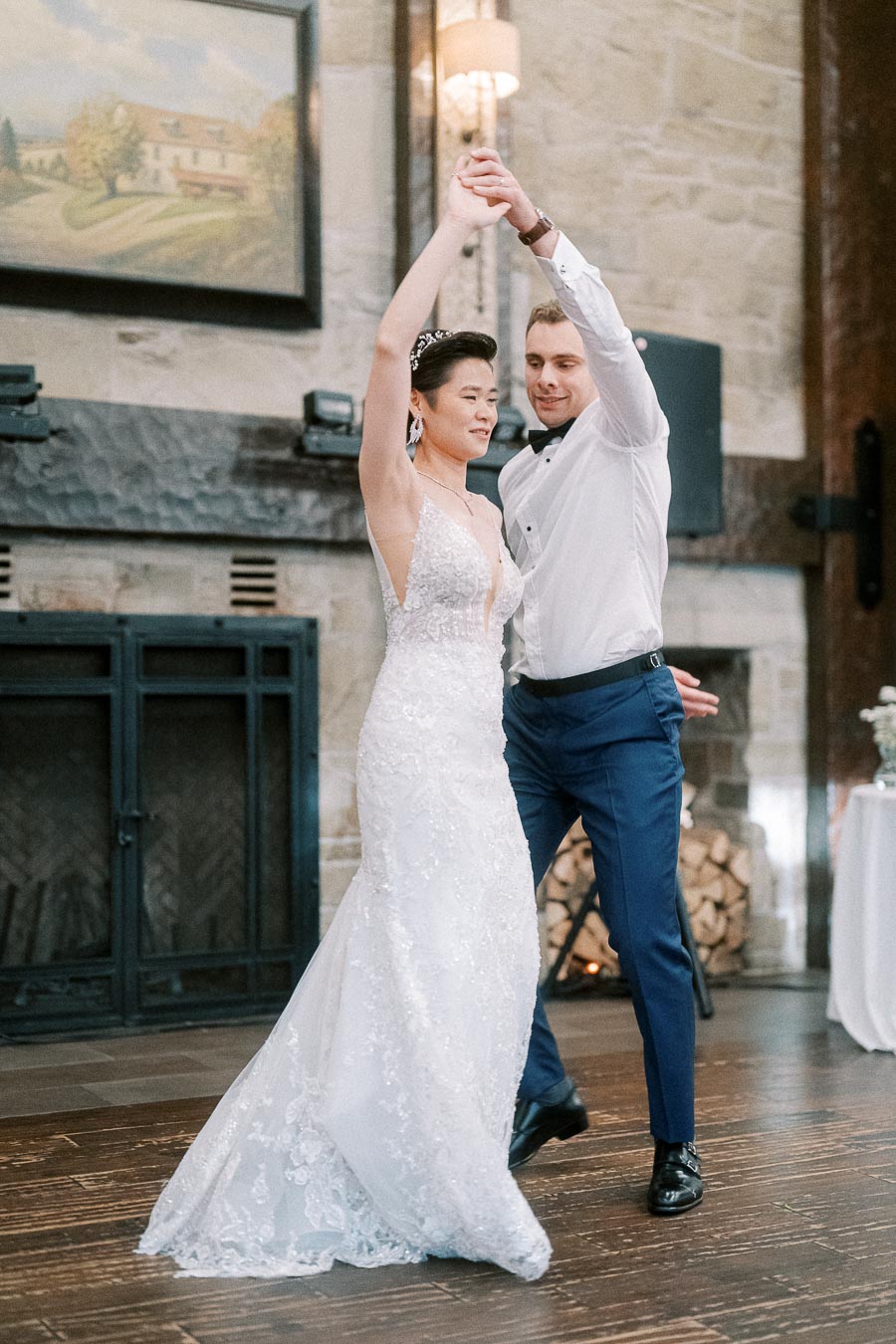 Bride and groom dancing elegantly in a rustic venue, showcasing a lace wedding dress and formal attire.