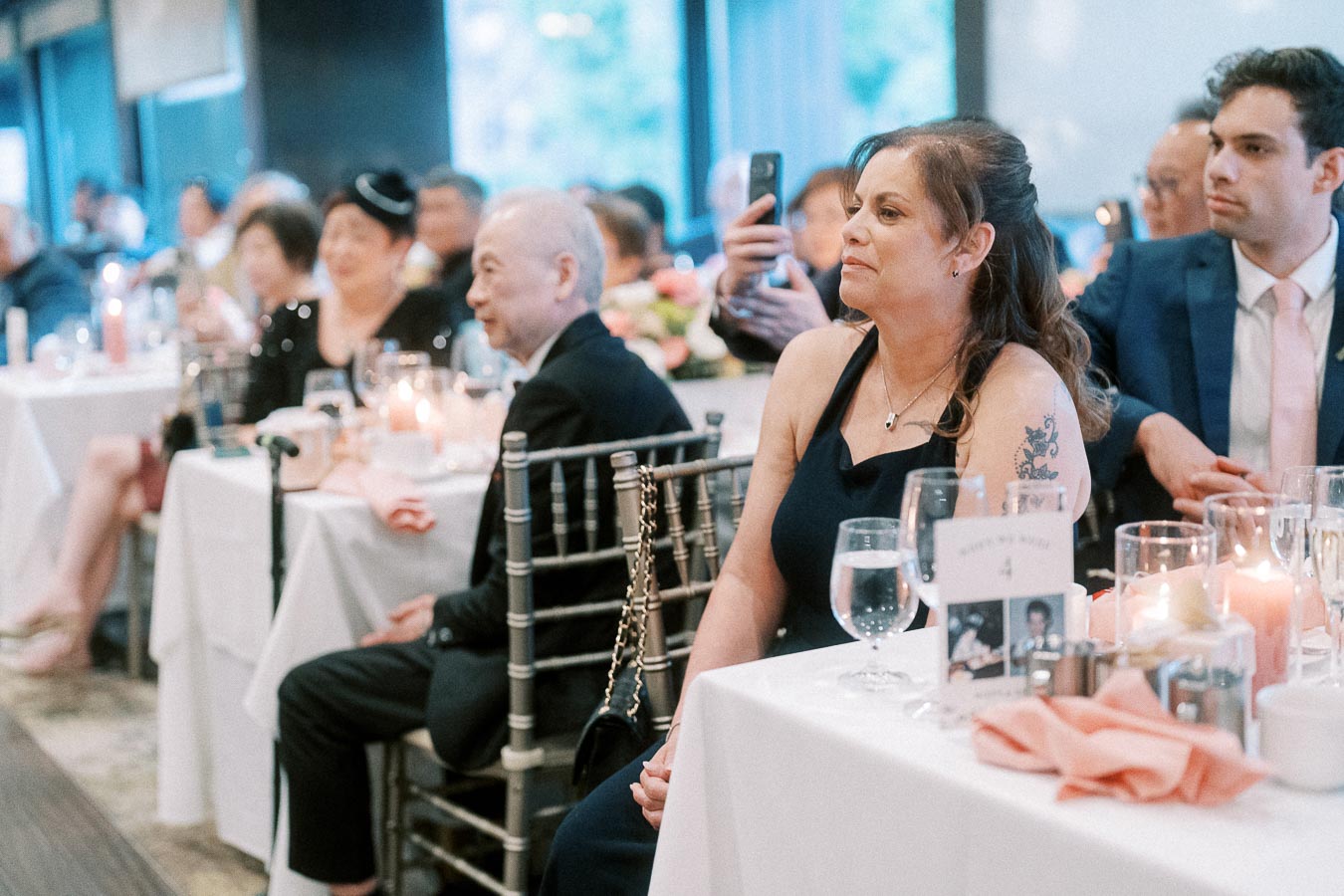 Guests seated attentively at an elegantly decorated event, featuring tables adorned with glasses, candles, and floral arrangements, creating a warm and sophisticated atmosphere.