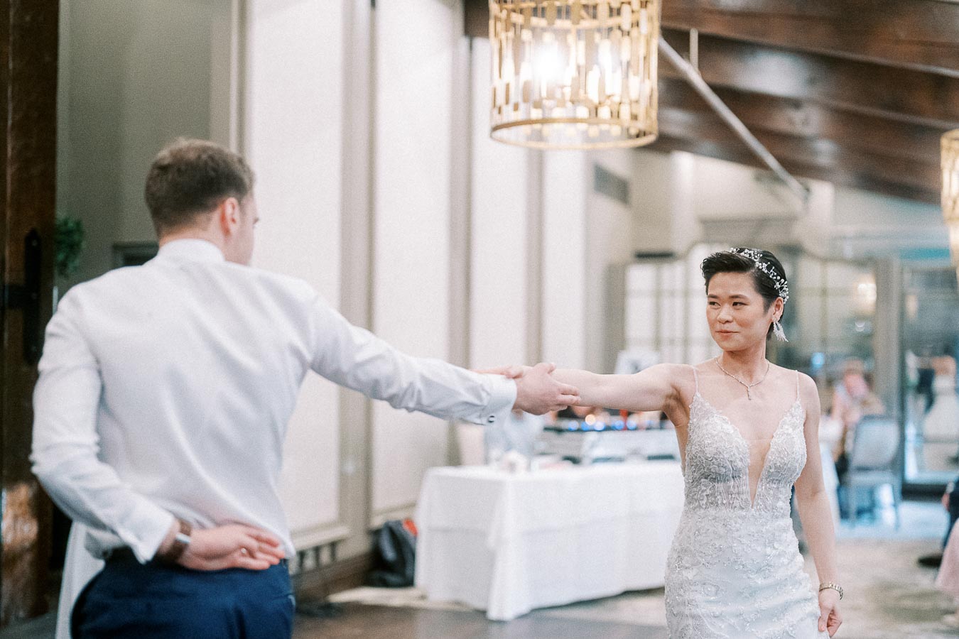 A couple elegantly dances in a decorated ballroom, with the bride in a stunning white gown featuring intricate lace and the groom in a classic white shirt, under warm ambient lighting.