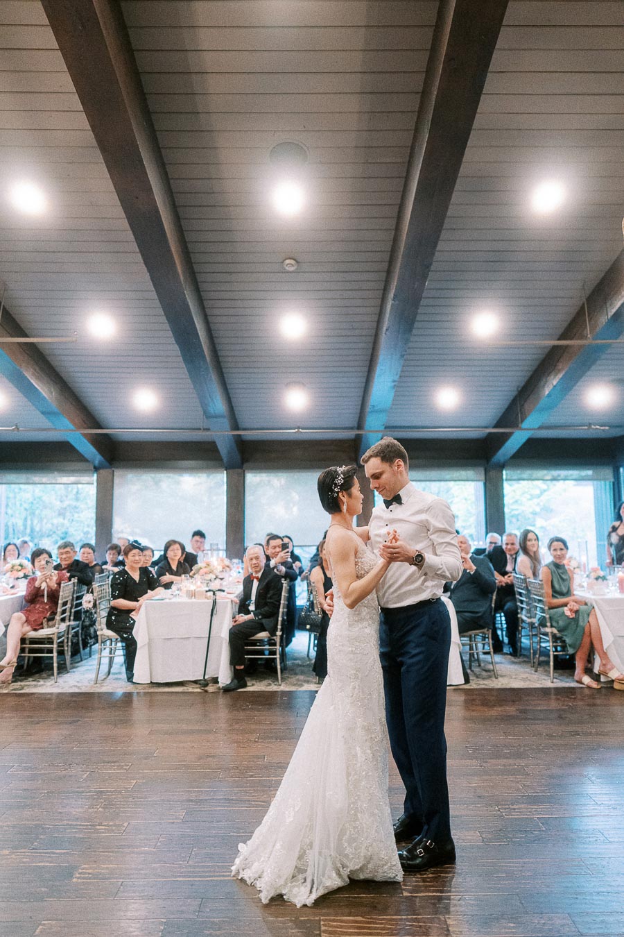 A bride and groom share their first dance in an elegantly decorated reception hall as guests watch and applaud, highlighting a joyful wedding celebration.