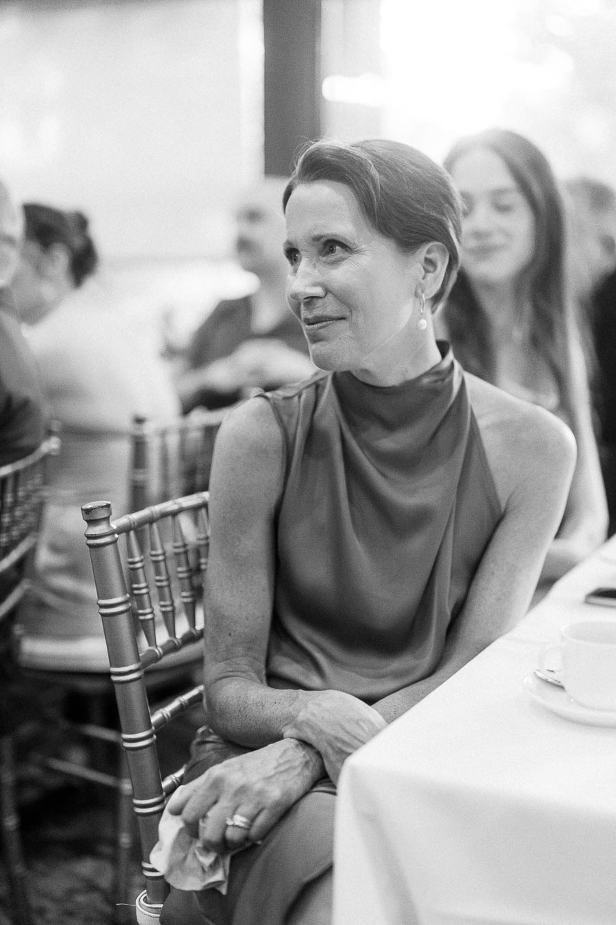 A woman in a formal sleeveless dress sits at a table during a social event, surrounded by other attendees.