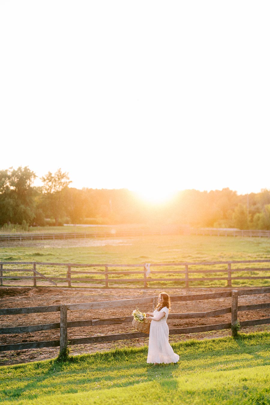Pregnant woman in a white dress holding flowers by a farm fence at sunset