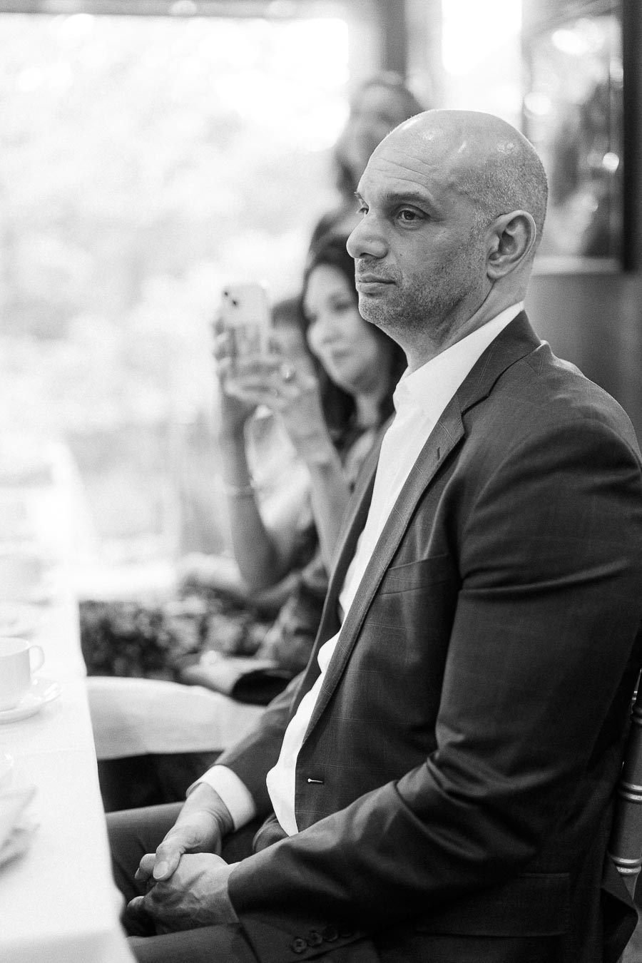Black and white photo of a thoughtful man in a suit sitting at a table during a formal event, with another person in the background capturing the moment on a smartphone.
