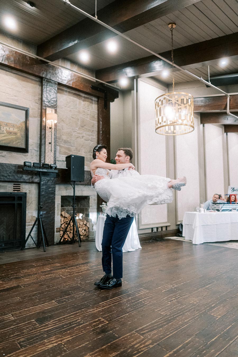 A groom lifting his bride in an elegant wedding venue with wooden floors and a stone fireplace, showcasing joyful celebration and dance.