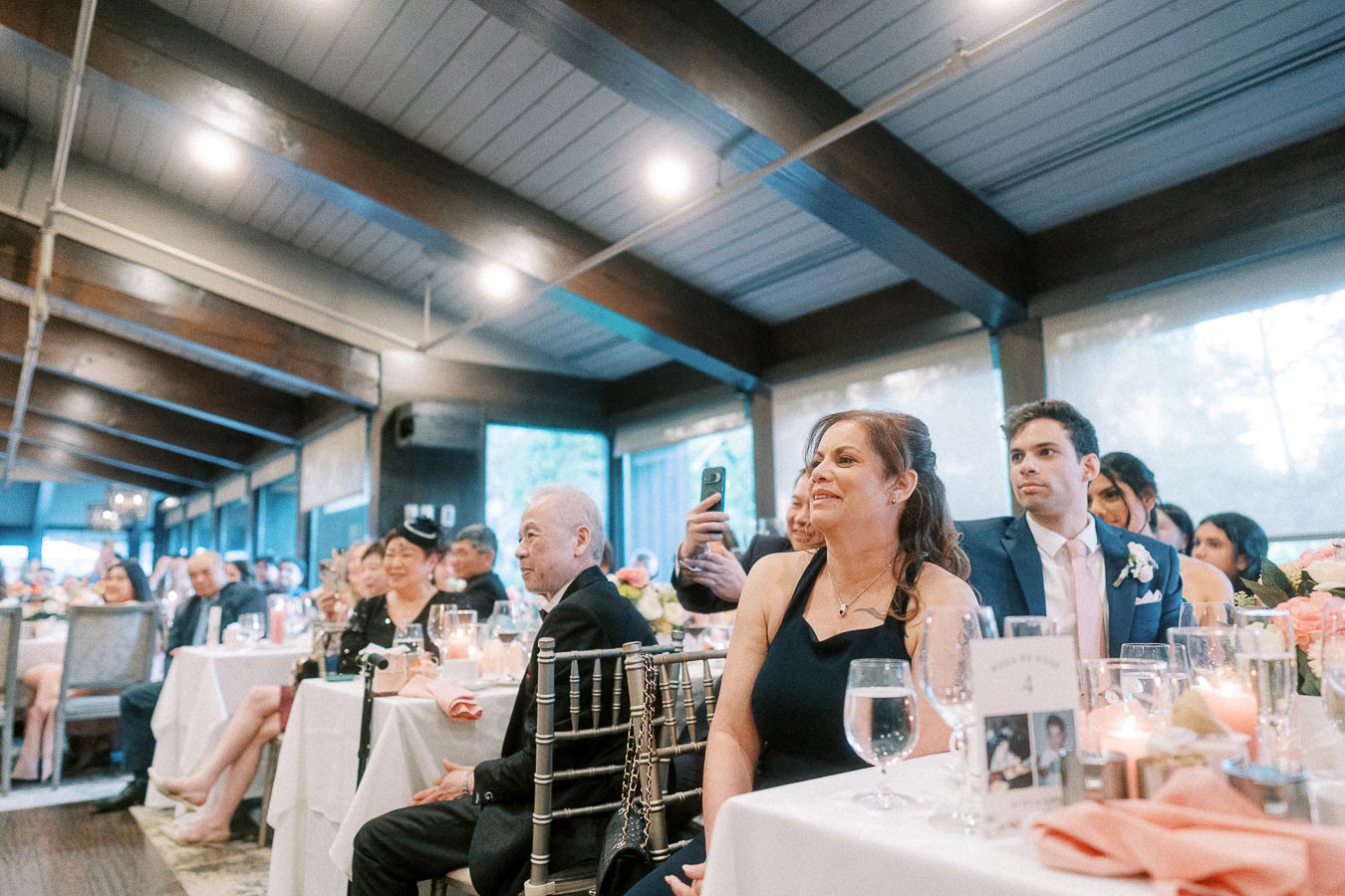 A diverse group of guests at a wedding reception, seated at elegantly decorated tables, attentively watching an event indoors.
