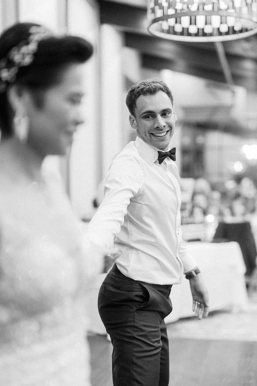 Black and white photo of a groom in a white shirt and bow tie smiling as he leads his bride during their wedding dance, capturing a joyful moment at the reception.