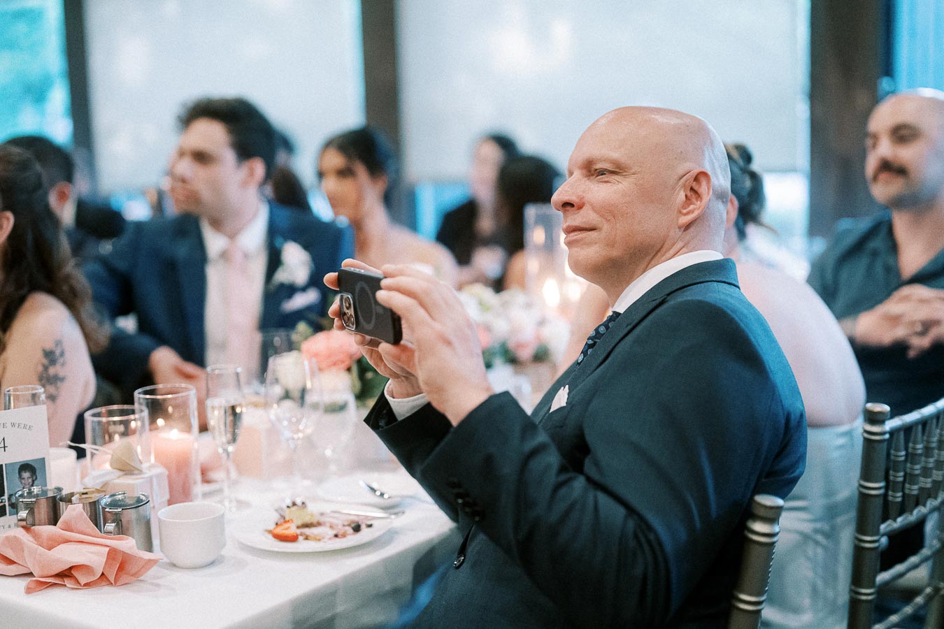 A man in a suit taking a photo with a smartphone at a wedding reception. The table is elegantly set with candles, floral centerpieces, and a variety of plates and glassware, while other guests sit in the background, engaging in conversation.