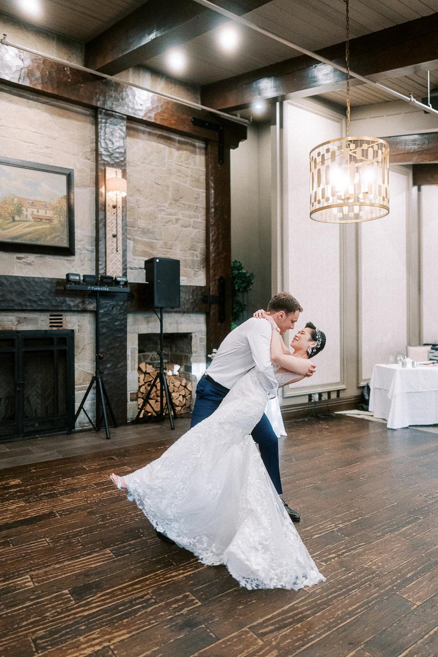 A newlywed couple dances elegantly in a rustic wedding venue, featuring a stone fireplace and wooden floors. The bride's lace gown flows gracefully as the groom dips her, creating a romantic and joyful wedding moment.