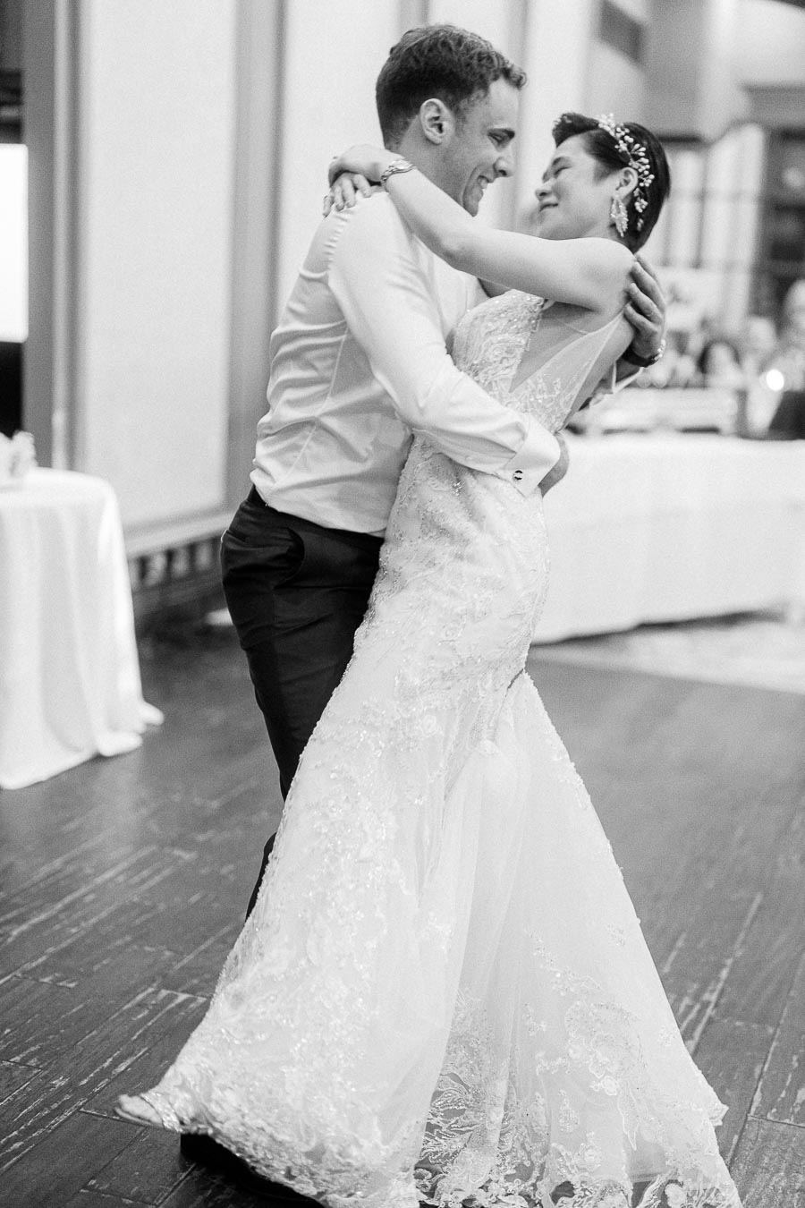 Black and white image of a joyful couple sharing a romantic dance at their wedding reception, with the bride wearing an elegant lace gown and the groom in a classic suit, captured on a wooden floor in a softly lit venue.