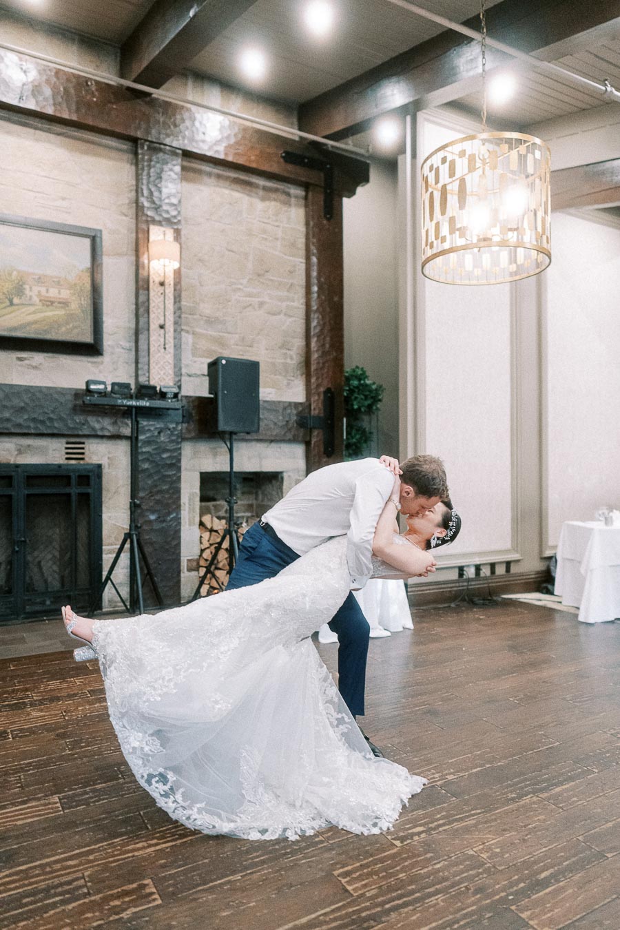A bride and groom share a romantic dance in an elegant reception venue, with the bride dressed in a lace gown and the groom in a white shirt and dark pants, beneath a modern chandelier.