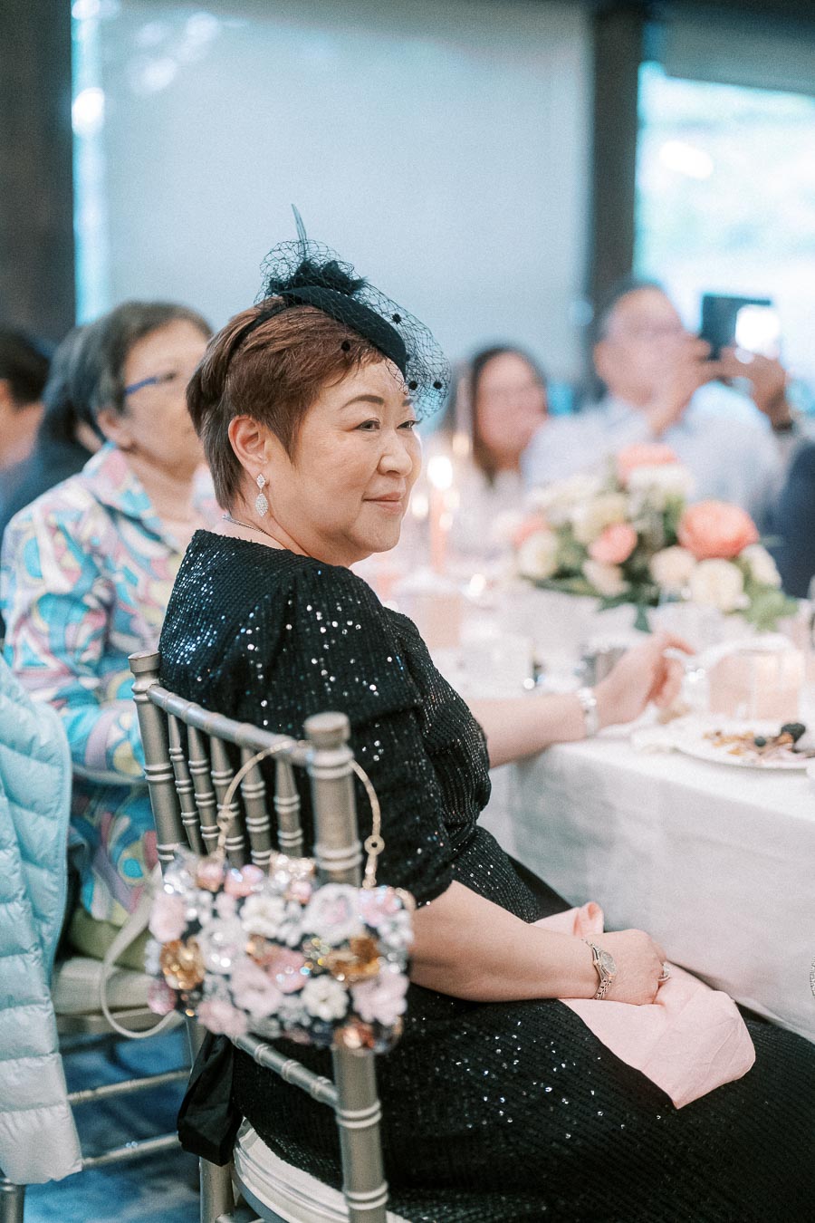 Elegant woman in a black sequin dress with a stylish fascinator, sitting at a beautifully decorated table during a formal event.