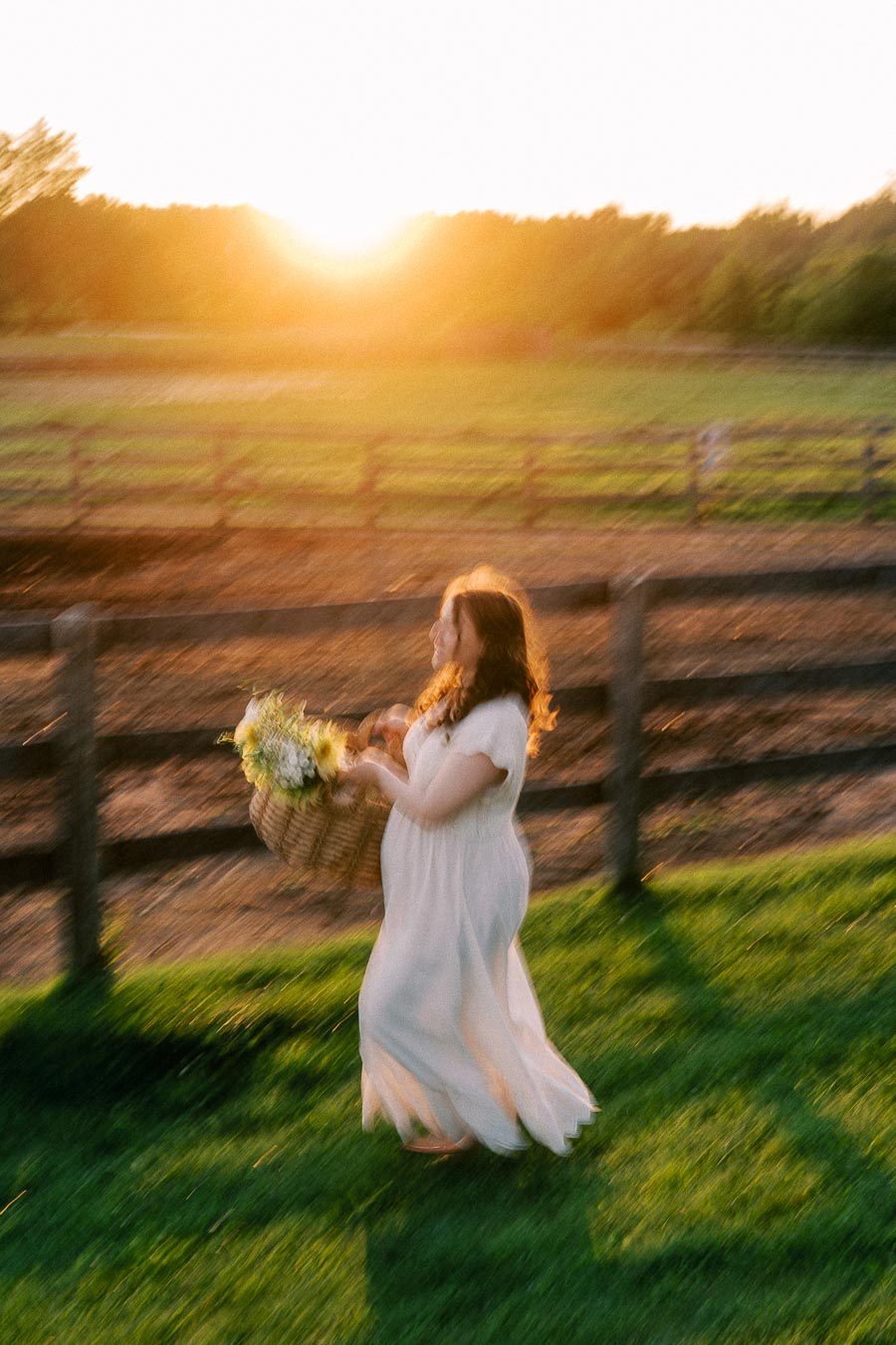 Pregnant woman in a white dress carrying flowers at a farm fence