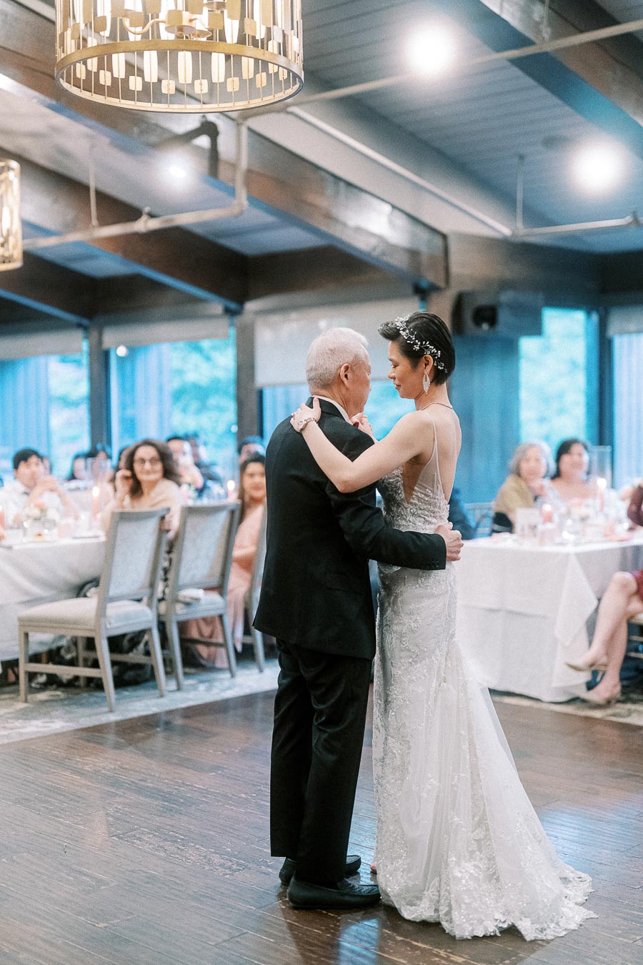 Father and bride share a heartfelt dance during a wedding reception, surrounded by seated guests, under soft and elegant lighting in a beautifully decorated venue.