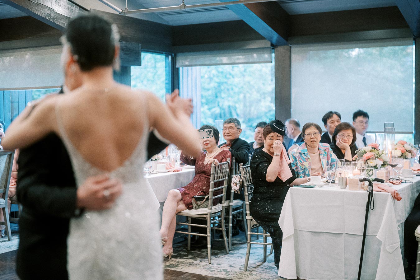 A bride and groom share a dance at a wedding reception, while seated guests watch and capture the moment on their phones. The elegant setting includes beautifully arranged tables and flower centerpieces, creating a warm and festive atmosphere.