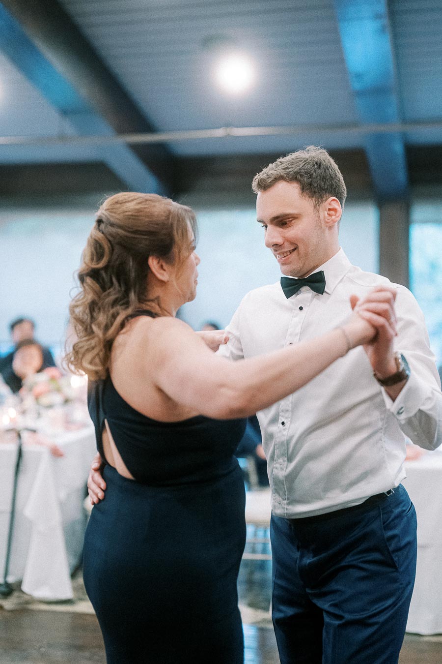 A couple elegantly dancing at a wedding reception, with the groom in a white dress shirt and black bow tie, and the woman in a sleeveless navy dress, surrounded by warmly lit tables in a beautifully decorated venue.