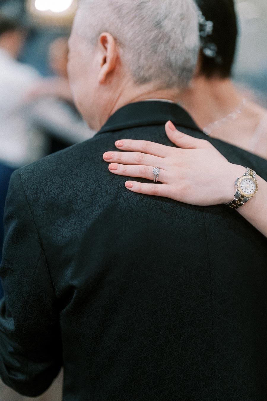 A close-up of a hand with a diamond ring and a gold watch resting on the shoulder of a man in a dark patterned suit. The image captures an intimate wedding dance moment.