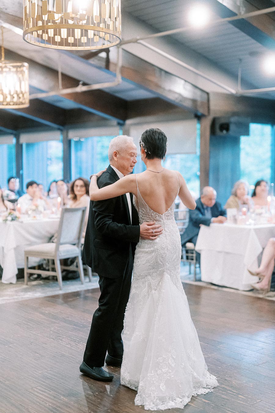 Father and bride sharing a heartfelt dance at a wedding reception, surrounded by elegantly decorated tables and attentive guests in a warmly lit venue.