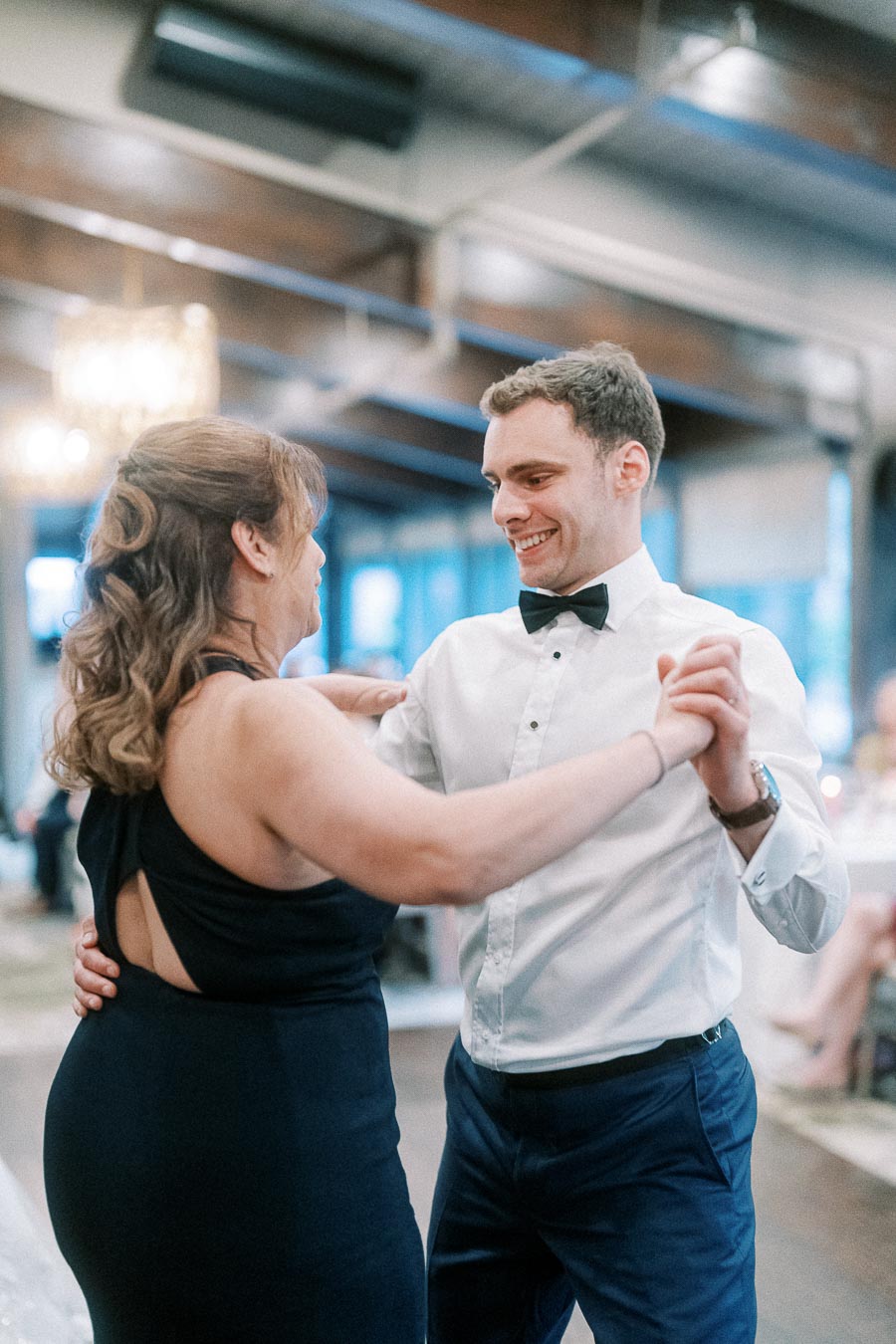 A couple joyfully dancing at an elegant indoor event, with the man wearing a white dress shirt and bow tie, and the woman in a black dress, highlighting a special moment of celebration and happiness.