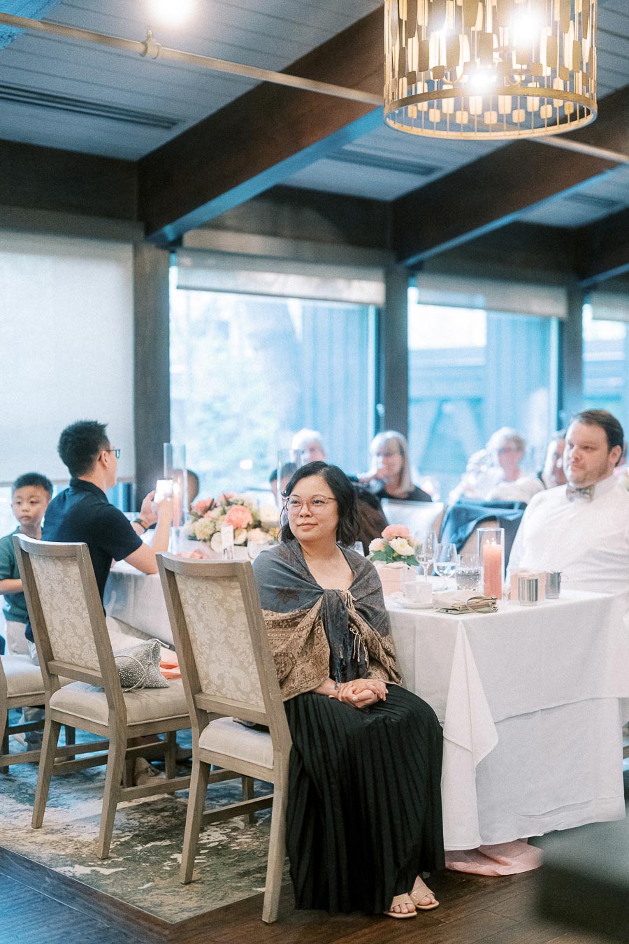A group of people seated at dining tables in a warmly lit restaurant, featuring elegant decor with flowers and candles on the tables.