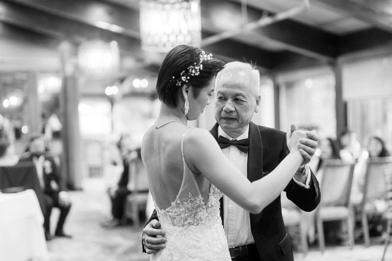 A bride in an elegant wedding gown shares a heartfelt dance with an older gentleman in a tuxedo at a wedding reception, surrounded by softly lit decor and seated guests in the background.