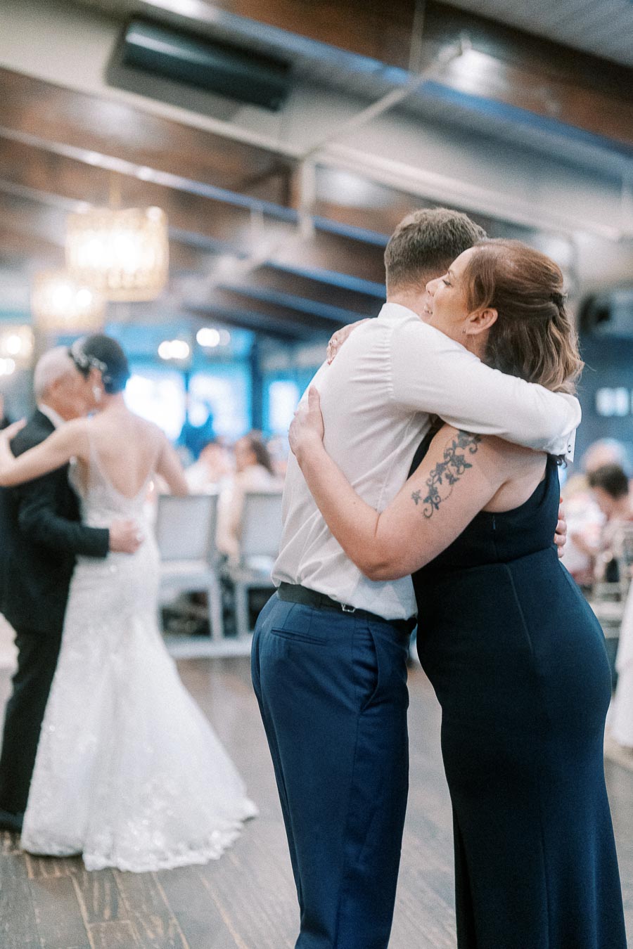 Couple embracing at a wedding reception, with another pair dancing in the background, under elegant chandeliers in a warmly lit venue.