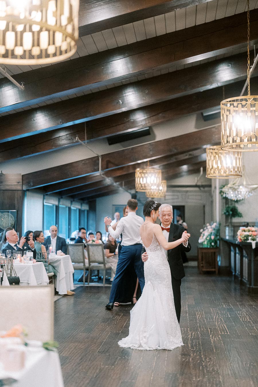 A bride in a white gown dances with an elderly man in a suit at a wedding reception in a warmly lit venue, surrounded by seated guests.