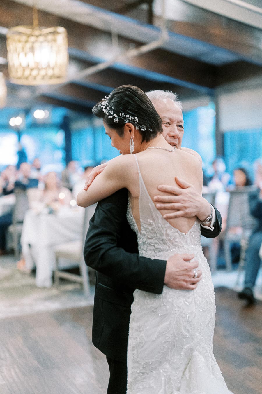 Father and bride sharing a heartfelt dance at wedding reception, indoor venue with guests in the background.