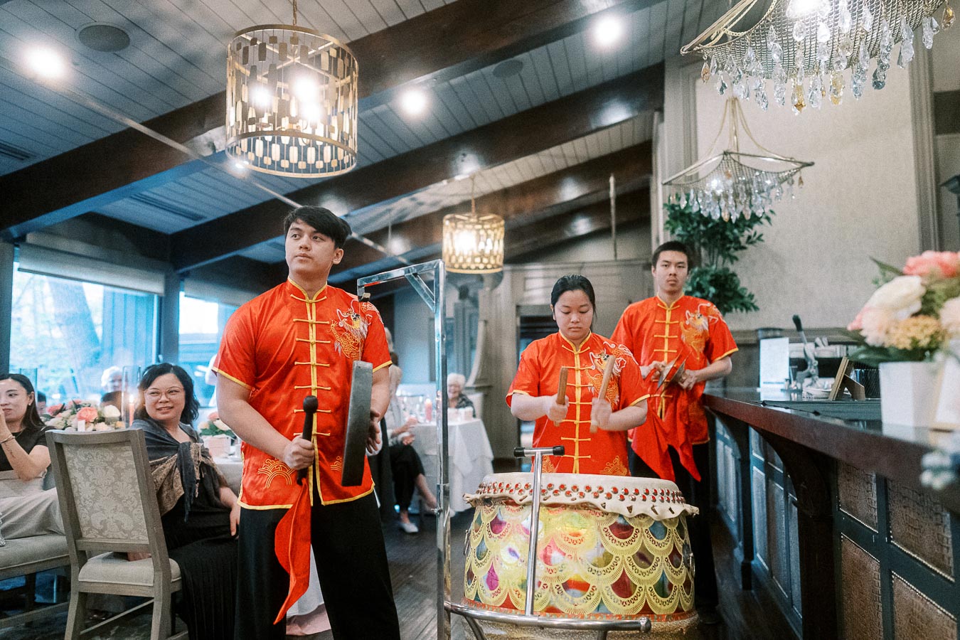 Performers in traditional red Chinese attire play a drum and gong at a festive indoor event, surrounded by elegantly decorated tables and chandeliers.