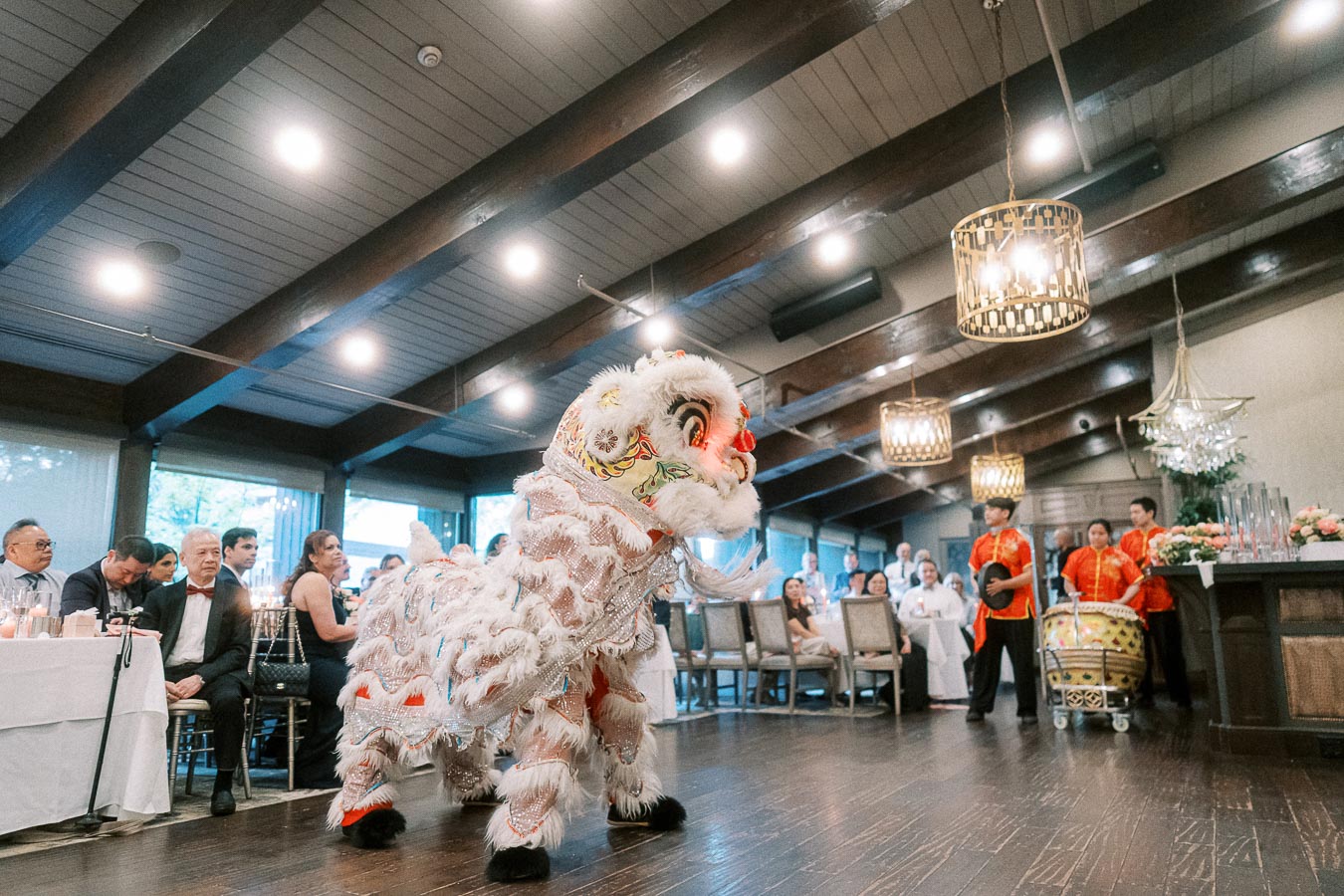 Lion dance performance indoors at a formal event, with a white furry costume, colorful embroidery, and musicians in red outfits playing traditional drums.