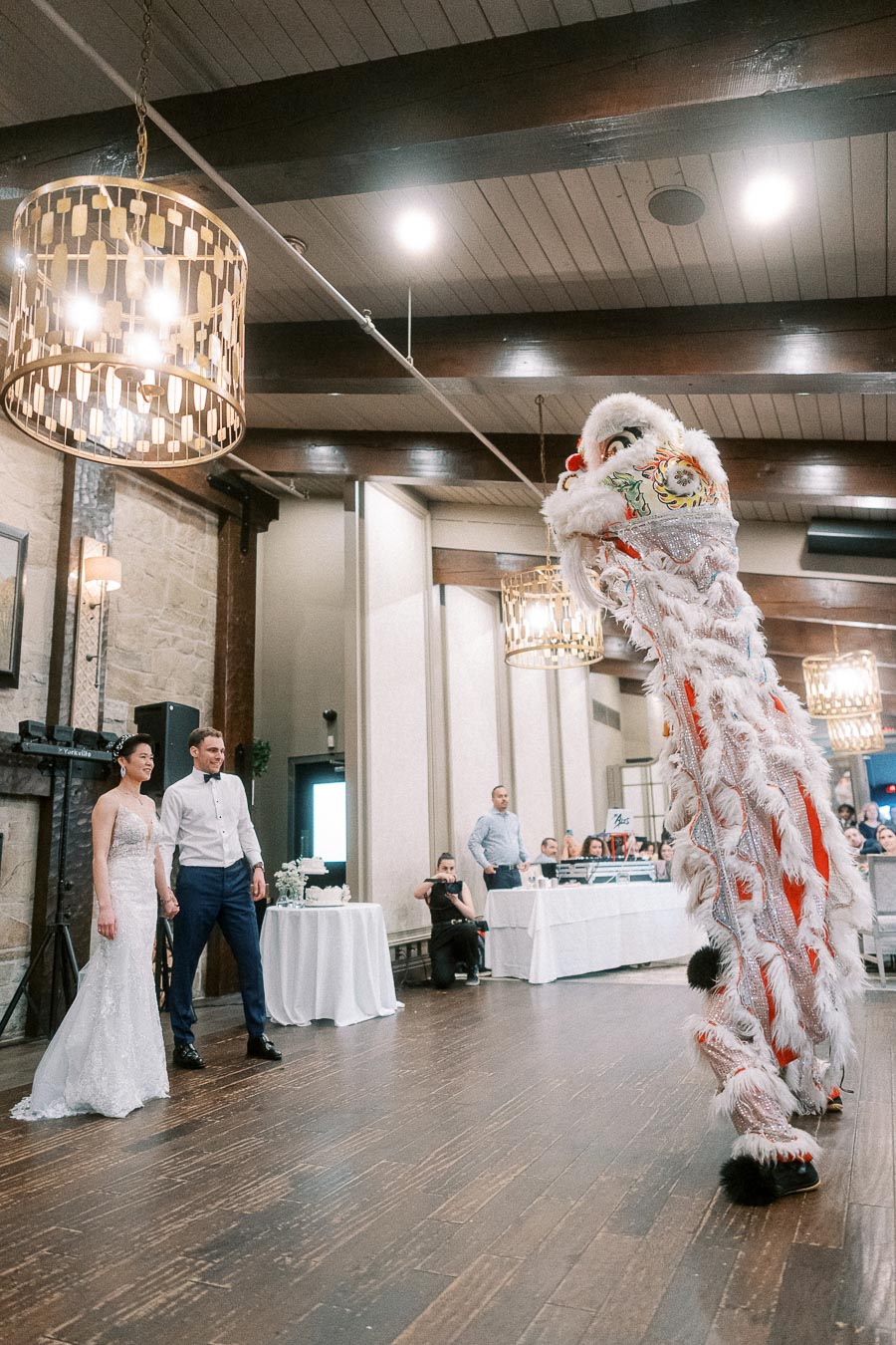 A bride and groom stand smiling as they watch a traditional Chinese lion dance performance at their wedding reception in an elegantly decorated venue.