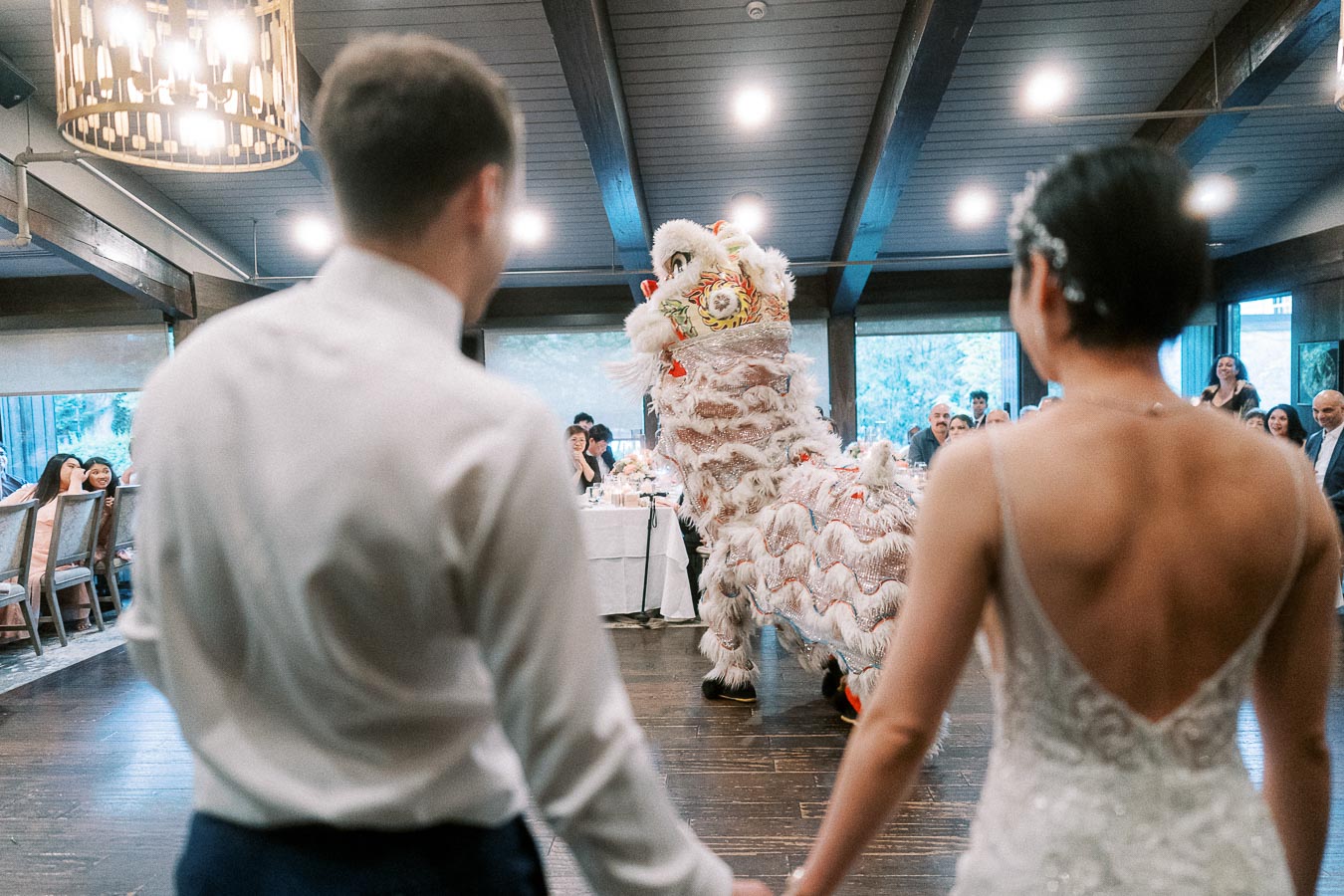 A couple watches a traditional lion dance performance at a wedding reception, surrounded by guests in a beautifully decorated venue with wooden beams and warm lighting.