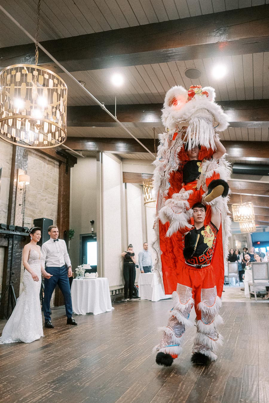 A traditional lion dance performance at a wedding reception, featuring performers in vibrant red and white costumes. The bride and groom watch excitedly in a beautifully decorated venue with wooden beams and elegant chandeliers.