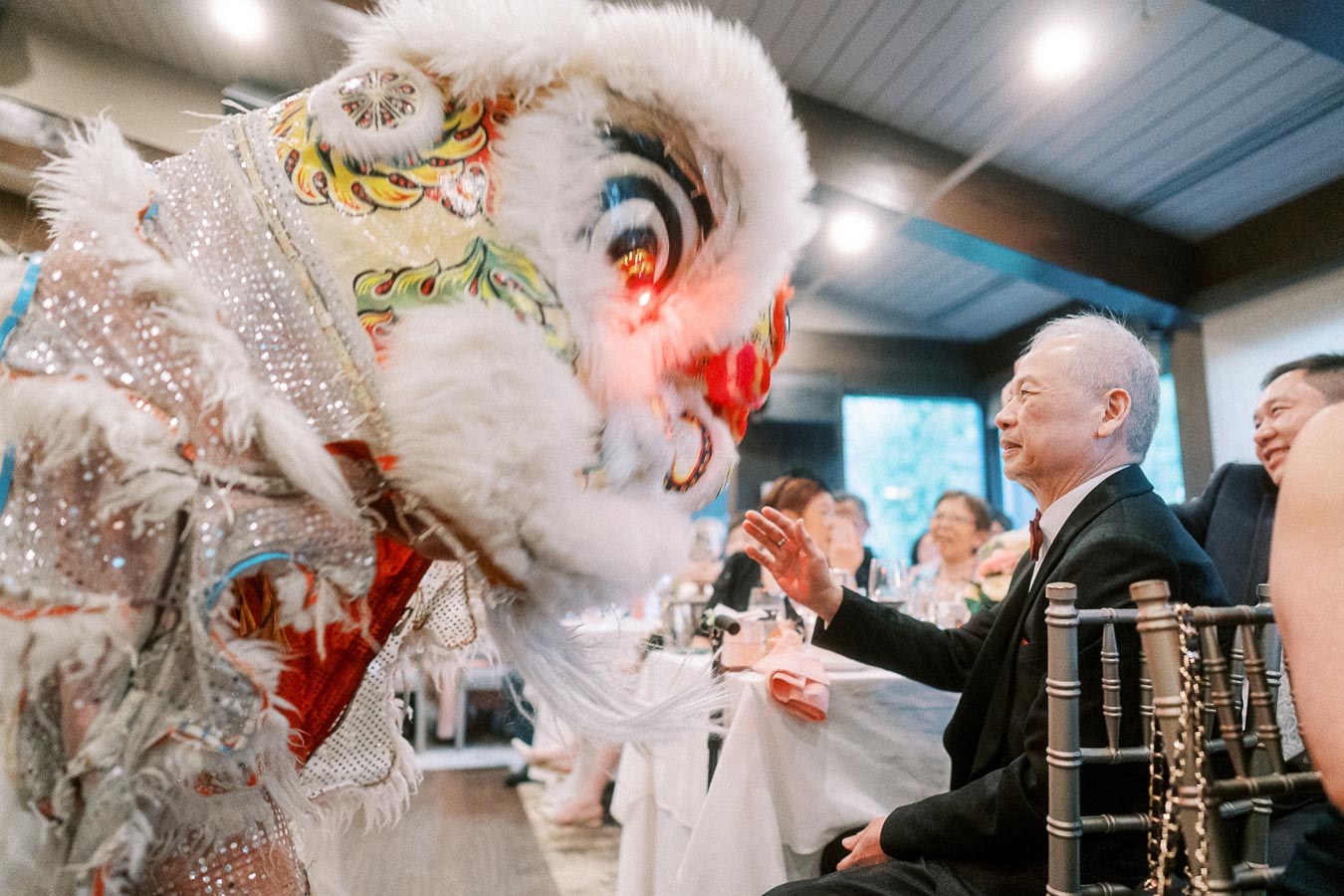 Elderly man enjoying a traditional Chinese lion dance at a festive indoor gathering, surrounded by seated guests in a well-lit venue.