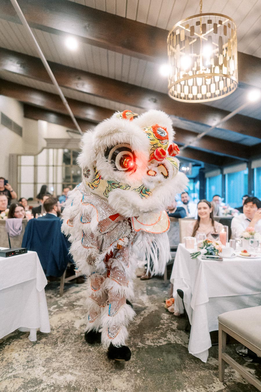 Lion dance performance at indoor event, featuring a colorful costume with intricate patterns and glowing eyes, surrounded by an audience of seated guests at decorated tables.
