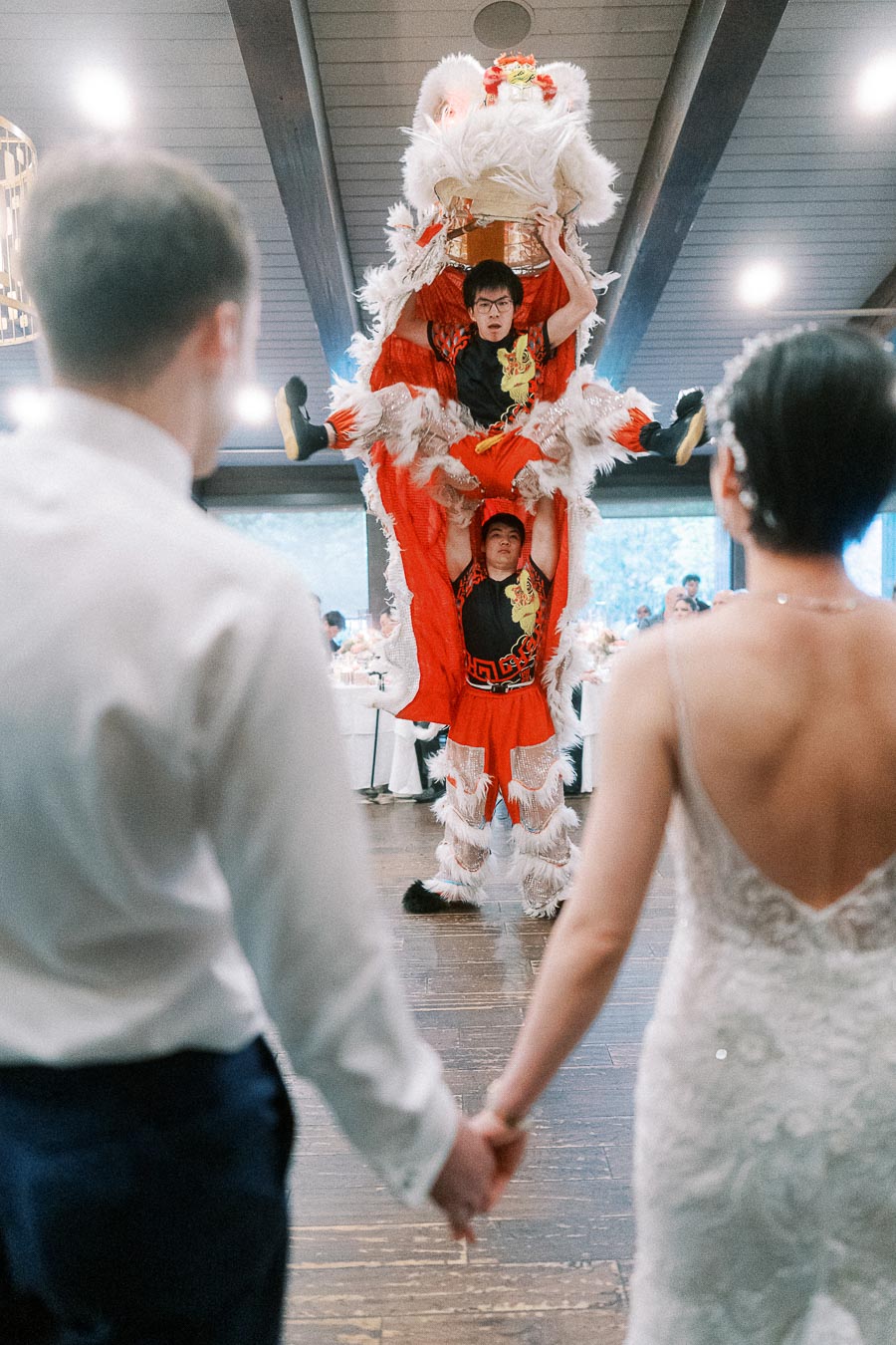 Bride and groom holding hands as performers entertain with a traditional red and white lion dance at a wedding reception.