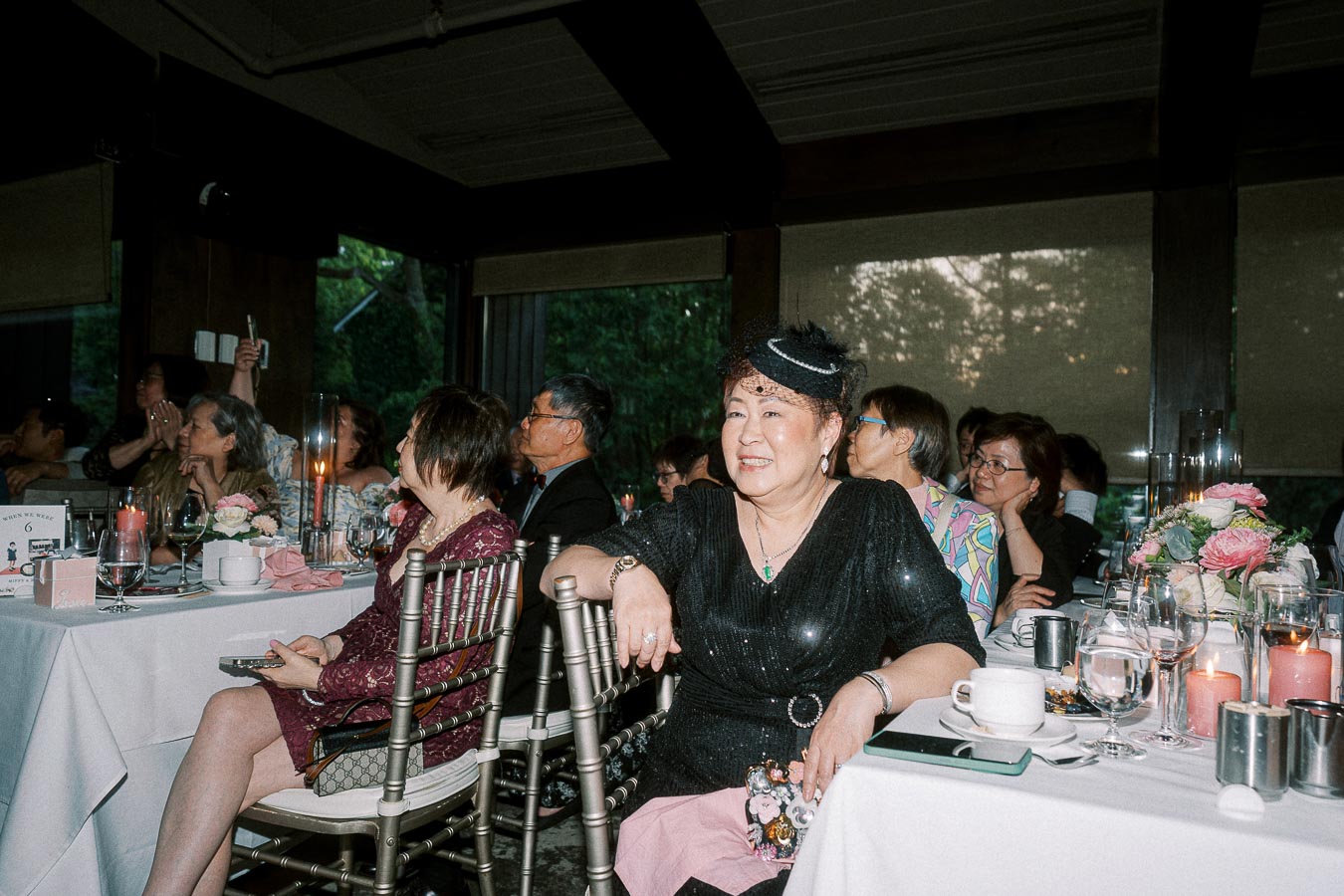 Guests enjoying a formal event at a restaurant, with a woman in a black outfit sitting by a decorated table with flowers and candles, surrounded by other attendees.