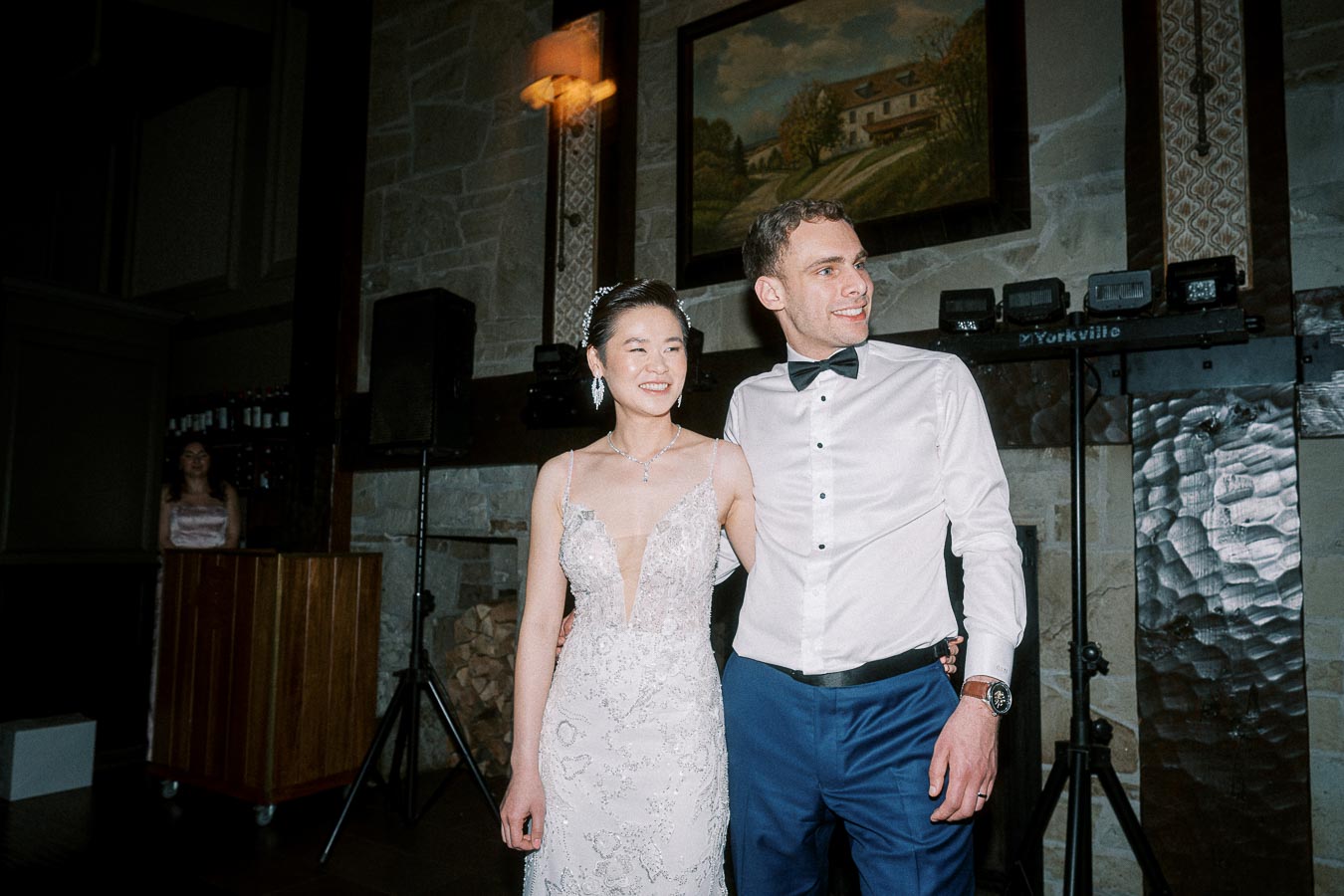 A happy couple posing together indoors, the woman in a detailed lace wedding dress and the man wearing a white shirt with a bow tie and blue pants, standing in a warmly lit setting with a rustic stone wall and a painting in the background.