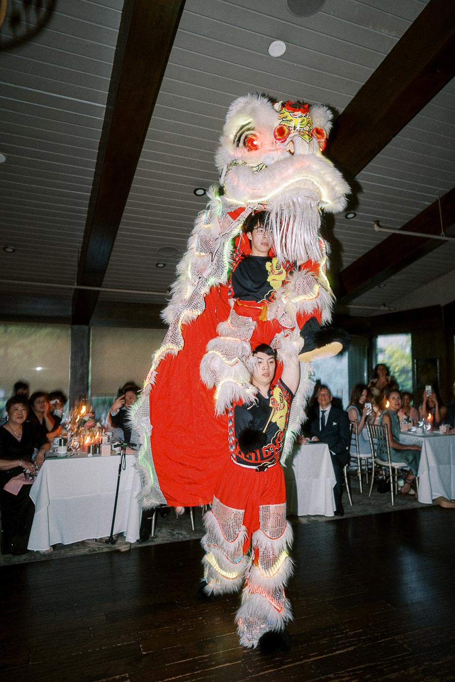Traditional Chinese lion dance performed indoors at a formal event, with guests seated around the room, showcasing vibrant red and white costumes and intricate detailing.