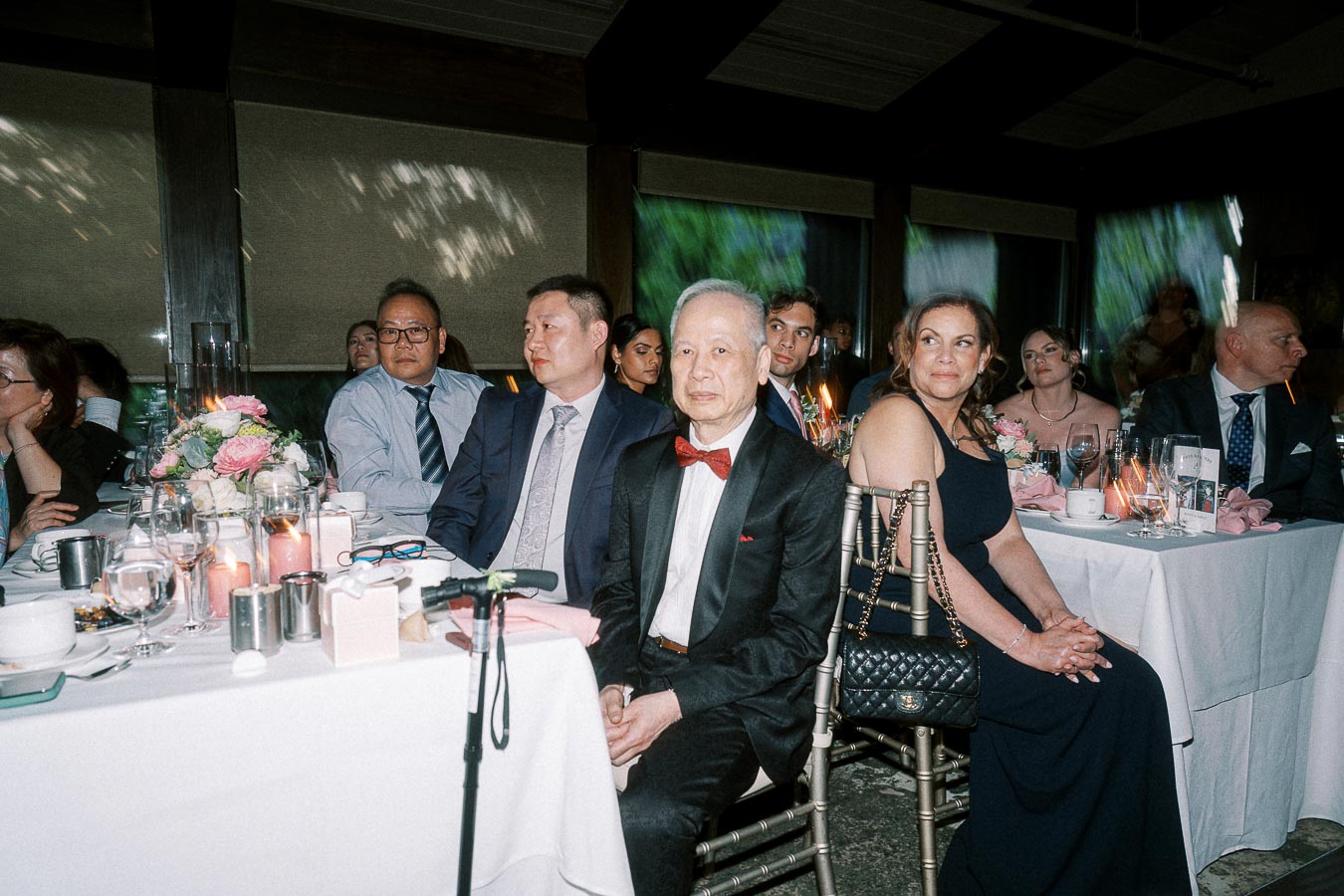 Guests attentively listening at an elegant formal event with decorated tables, floral centerpieces, and candlelight ambiance.