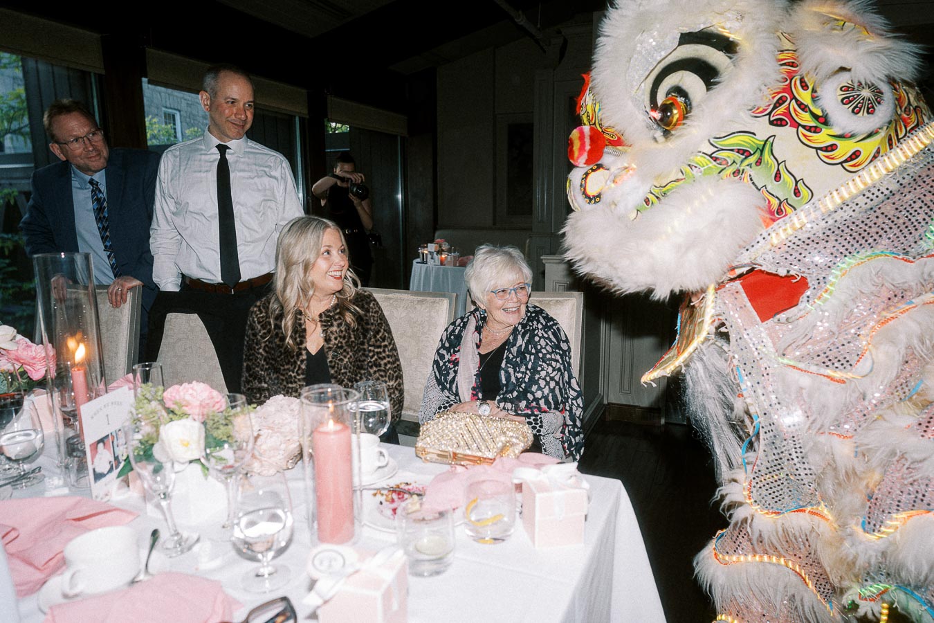 A vibrant Chinese lion dance performance entertains guests at an elegantly decorated dinner table. The scene highlights the colorful lion costume and guests' joyful reactions, surrounded by pink floral arrangements and candles, creating a festive atmosphere.