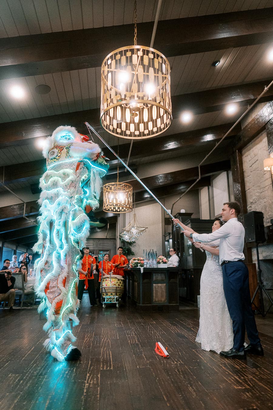 A bride and groom participate in a vibrant lion dance performance inside a decorated venue, with the illuminated lion costume captivating the guests.