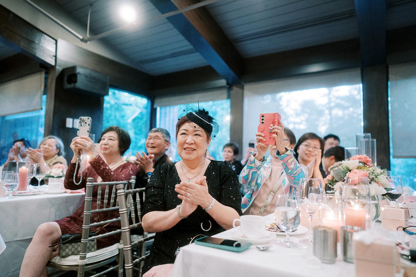 Group of people smiling and clapping at an indoor event, with some capturing the moment on smartphones.